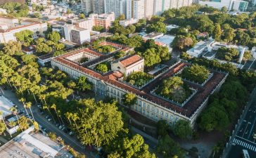 Vista aérea de um campus universitário histórico, cercado por áreas verdes, no Rio de Janeiro.
