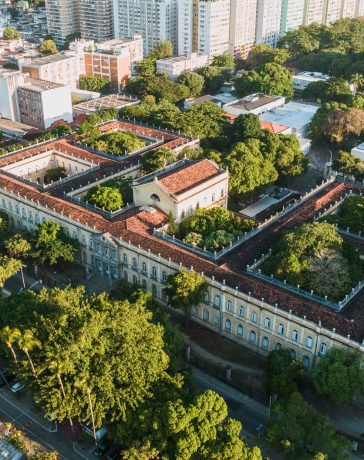 Vista aérea de um campus universitário histórico, cercado por áreas verdes, no Rio de Janeiro.