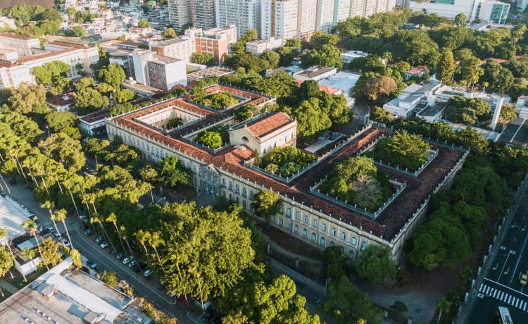 Vista aérea de um campus universitário histórico, cercado por áreas verdes, no Rio de Janeiro.