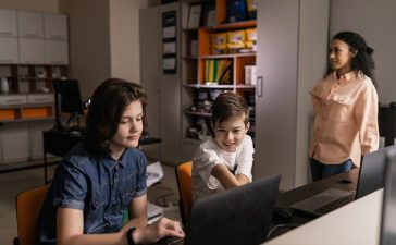 Estudantes usam laptops em sala de aula enquanto um professor acompanha a atividade.