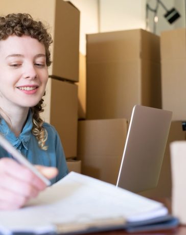 Empreendedora sorridente trabalha com um laptop enquanto organiza pacotes de sua pequena empresa.