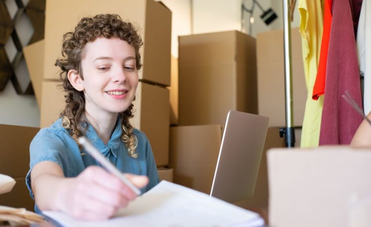Empreendedora sorridente trabalha com um laptop enquanto organiza pacotes de sua pequena empresa.