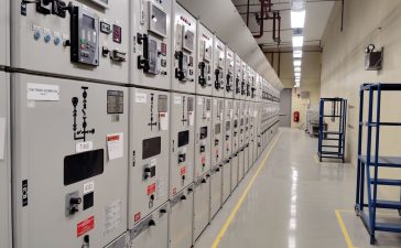 Sala de controle elétrico com uma longa fileira de painéis industriais iluminados.