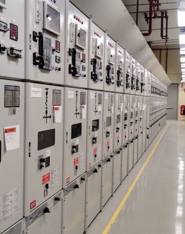 Sala de controle elétrico com uma longa fileira de painéis industriais iluminados.