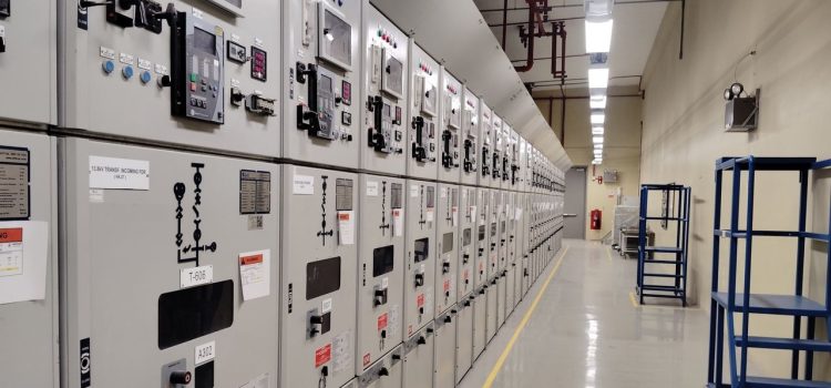 Sala de controle elétrico com uma longa fileira de painéis industriais iluminados.