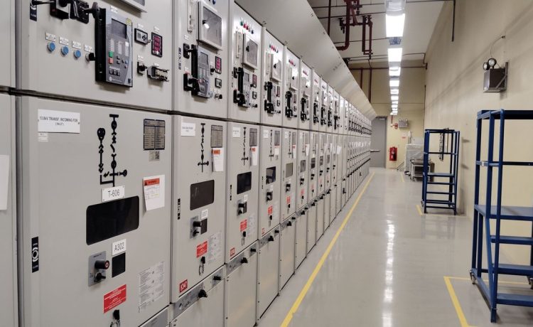 Sala de controle elétrico com uma longa fileira de painéis industriais iluminados.