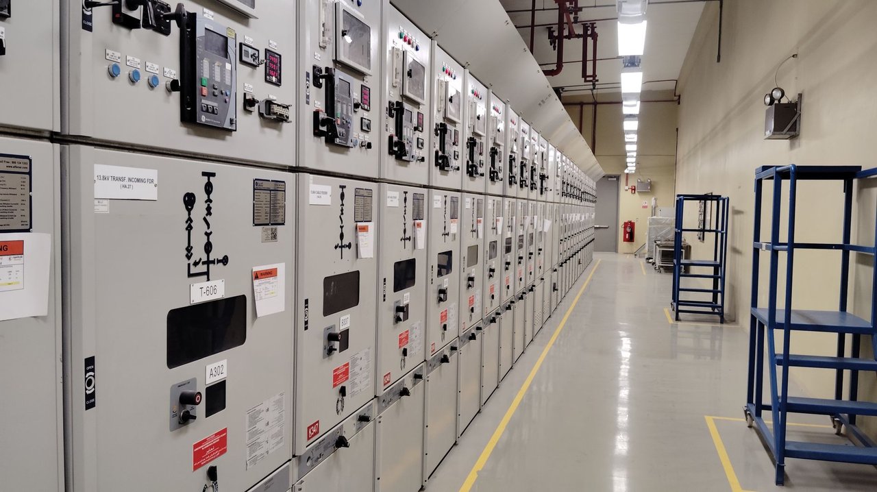 Sala de controle elétrico com uma longa fileira de painéis industriais iluminados.
